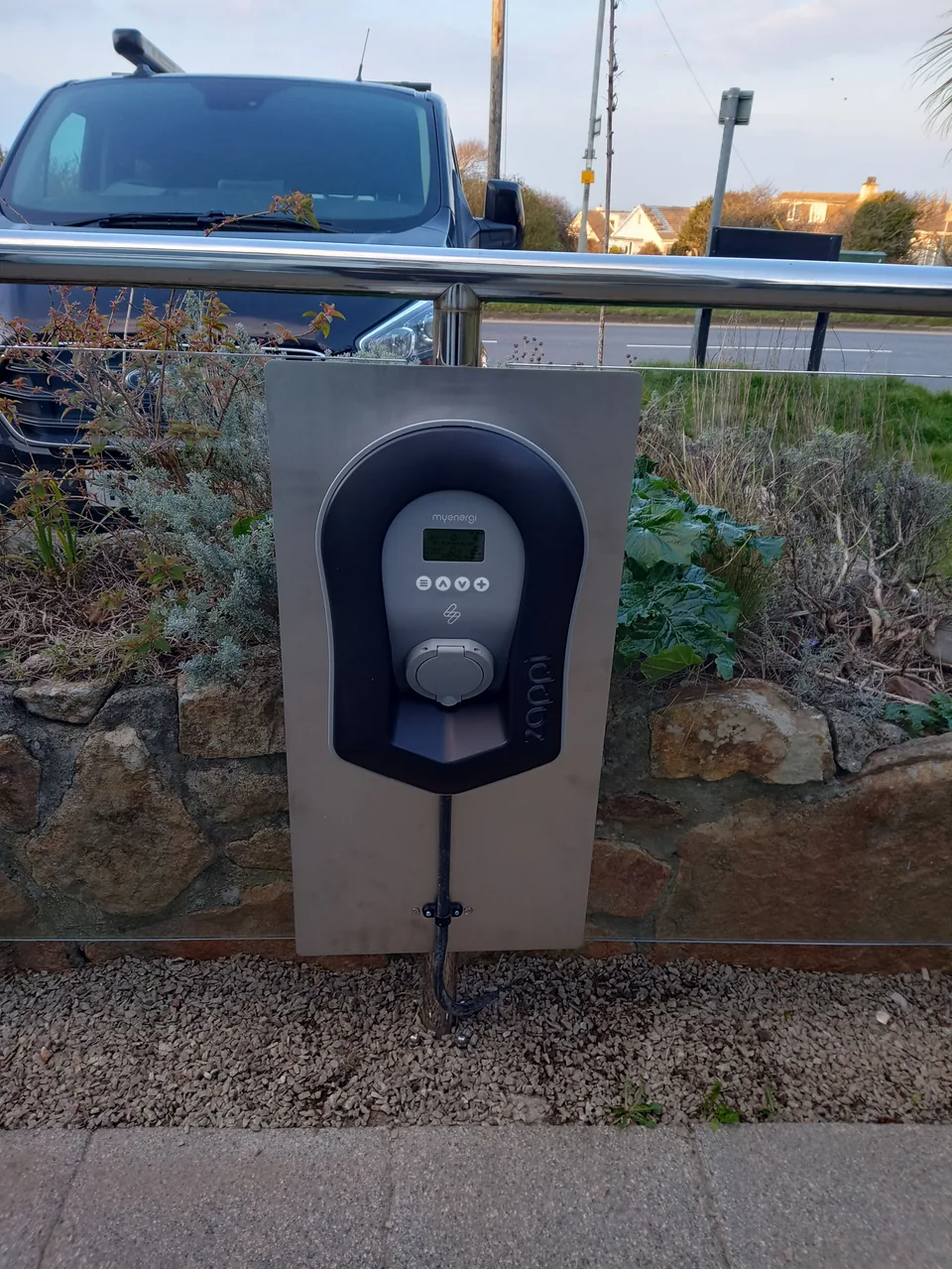 Zappi EV charger fitted to a rectangular stainless steel backplate beside a fence with glazed panels